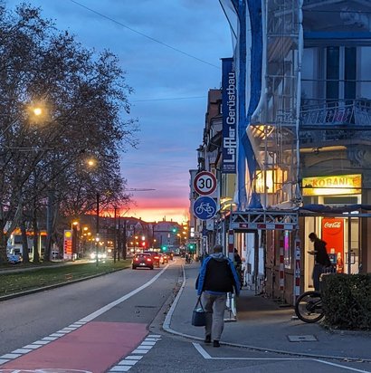 Eine beleuchtete Straße in der Abenddämmerung; im Vordergrund ein roter Fahrradweg, rechts daneben ein Laden mit rotem Cola-Automaten im Eingang.